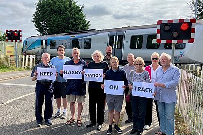 Lib Dem campaigners holding up signs saying 'Keep Haxby Station On Track', standing in front of a level crossing with a train passing behind