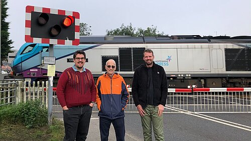 Councillors Pearson, Cuthbertson and Hollyer standing in front of a level crossing as a Transpennine Nova 3 passes behind them